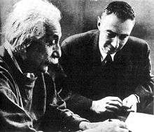 Einstein writing at a desk. Oppenheimer sits beside him, looking on.