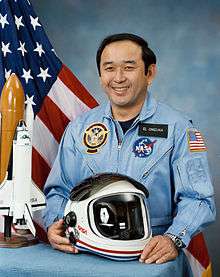 An image of LTC Onizuka with a model of the Challenger shuttle and astronaut helmet on a desk in front of him. The United States Flag, and a smoky blue backdrop in the background.