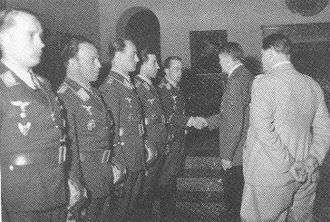 Five men all wearing military uniforms and decorations standing in row. The man on the far right is shaking hands with another man whose back is facing the camera. Another man is standing behind the men shaking hands.