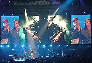 A large screen with the silhouette of a man, seated with two pistols, is in the center. Surrounding it are screens showing Eminem, who is to the right of Rihanna. The latter is holding a microphone and has short red hair.