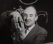 Photograph of the head, shoulders, and hands of a man standing in front of a blackboard. The man is smiling and staring at this hands, which are at to the left of his face in the photograph; he is apparently using his hands to explain something. The fingers of his left hand are pointing upwards. His right hand is above his left, with its fingertips pointing downwards and touching the matching fingertips of his left hand. The two hands thus form an enclosure. He is dressed in a tweed jacket with a necktie. There is a diagram chalked on the blackboard.