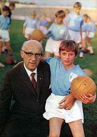 A photograph of an older man with a boy sitting in his lap. The older man, to the left, is bald, wearing glasses and wearing a dark brown business suit with a white shirt and a red tie underneath. The boy is blonde and wears a light blue football shirt and white shorts, with a leather football in his left hand. Several other boys can be seen with similar outfits in the background.