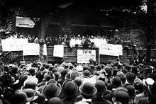 Black and white photograph of a speaker rallying a large crowd. In front of the stage, facing the audience, are several signs, in various languages, displaying demands.