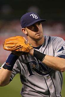 A man in a gray and blue baseball uniform with the letters "TB" on his cap holds a baseball in his glove, preparing to throw.