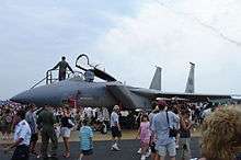 A jet sits on the tarmac while a crowd surrounds it. People are seen climbing into the jet's cockpit.