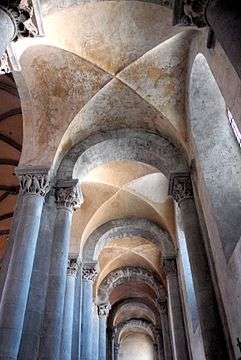 A narrow space with grey columns with ornate capitals supporting a plastered cross vault without ribs.