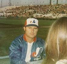 A man in a blue warm-up jacket and red, white, and blue baseball cap looks at the camera. A person with brown hair stands in the right-hand foreground.