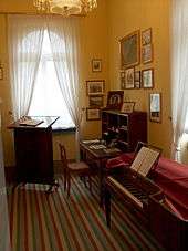 room furnished in early nineteenth century style, with striped runner on floor; walls painted peach and wall on right hung with small pictures; in the right foreground a square piano, in left rear before a window a reading stand, in right rear a desk, all in dark wood