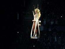 A blond female performer floating in a silver platform above the audience. She is wearing a silver outfit and a white mask.