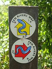 A blue eel symbol on yellow arrow on a white circle, and a blue eel symbol on red arrow on a white circle, both nailed to a wooden post, with a green leafy background.