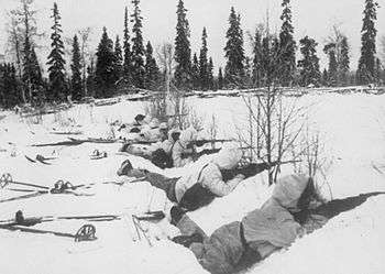 A group of soldiers with snowsuits and skies lies on the snow, guns pointing to the right.