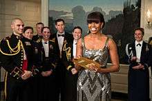 An African-American female in her late forties is seen wearing a silver dress and holding a golden envelope. Several men and women wearing black military garb are standing behind her.