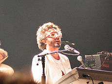A man with glasses in a white suit singing to a microphone and playing the keyboards.