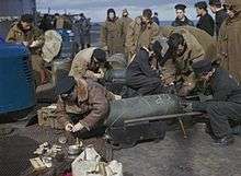 Colour photograph of men wearing military uniform crouching over bombs on the flight deck of an aircraft carrier