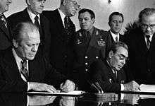 Two men in suits are seated, each signing a document in front of them. Six men, one in a military uniform, stand behind them.