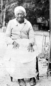 Black and white photo of an elderly African American woman looking at the viewer, wearing turn of the 20th century work clothes, a do-rag, and seated with her hands on her knees