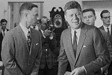Black-and-white film screenshot showing the main character on the left looking towards another man, President Kennedy, (voiced by actor Jed Gillin), on the right. Kennedy is smiling and looking to his left. In the background several men are looking in different directions and one is aiming a camera.