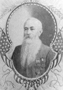 Head and torso of an older white man with a long flowing beard wearing a jacket with four medals hanging from ribbons pinned to the left breast. The portrait is surrounded by a decorative frame and illustrations of American flags and laurel wreaths.