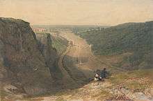 Two people on a cliff top look along the length of a river with wide muddy banks which snakes through a gorge towards a the distant city of Bristol. A line of small boats sail along the river.