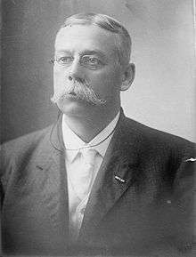Head and shoulders of a white man with a mustache and pince-nez glasses, wearing a dark suit coat over a light-colored vest, shirt, and tie.