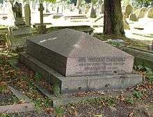 A red granite gravestone geometrically similar to the roof of a house, surrounded by other headstones