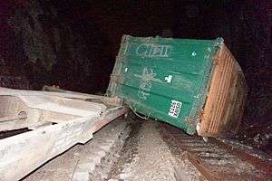 A large green metallic container lying on its side across some railroad tracks, one corner resting on a concrete structure on the left, seen at night.