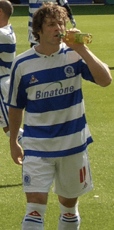 Young white man with dark curly hair wearing blue-and-white sports kit leaves a sports field.