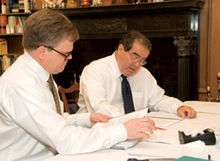 Two men in shirtsleeves work at a table, there are quantities of paper in front of them..
