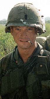 A man is at the center of the image looking at the camera. He is dressed in Vietnam War–era military attire including a vest and helmet. He has a cigarette sitting on his lips and is wearing a backpack.