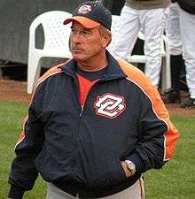 A man walking with a hand in his pocket. He is wearing a black and orange baseball cap and jacket.