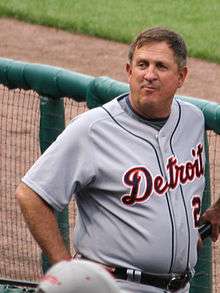 An older man standing in a baseball dugout wearing a grey uniform with "DETROIT" on the chest.