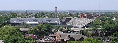 An elevated view of several buildings and the trees surrounding them. A red brick building with a sloped roof is in the foreground, and a large white football stadium is just behind it, taking up much of the center of the picture. Beyond the stadium, there is a red brick smokestack near the center of the picture, the red brick Tech Tower building on the left side bearing white letters that spell "TECH", and the red brick physics building on the right side. In the background there is a white domed building. All around these buildings are green-leafed oak trees. An overcast, light blue sky takes up the top third of the picture.