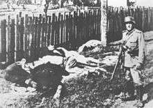 black and white photograph of several corpses in civilian clothes lying on the ground, with a male in German uniform standing near them