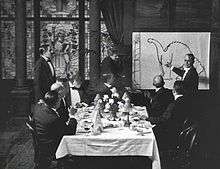 A black and white film still.  A group of men sit around a dining table in the center.  To the right, a man stands by and gestures at a large drawing of a dinosaur.