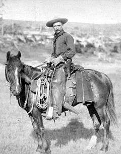 A black-and-white photograph of a cowboy posing on a horse with a lasso and rifle visible attached to the saddle