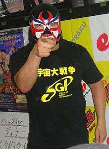 A color photograph of a Japanese man in a black mask with a white front and red and blue markings pointing at the camera
