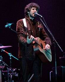 A man with long, bushy dark brown hair and a short beard playing acoustic guitar while singing into a microphone and looking to his right, away from the camera. He is seen from the knees upwards and is wearing a brown jacket with a red and blue embroidered pattern on the right shoulder.
