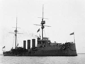 A large dark gray warship sits motionless in the water; it has four tall, thin smoke stacks closely arranged in the middle of the ship with two tall masts on either end.
