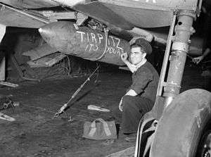 Black and white photograph of a man wearing military uniform crouching under an aircraft. He is holding a piece of chalk in his hand, and is posing next to a bomb which is fixed to the bottom of the aircraft on which "Tirpitz it's yours" has been written.