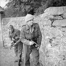 Two men in uniforms advance behind a wall carrying weapons