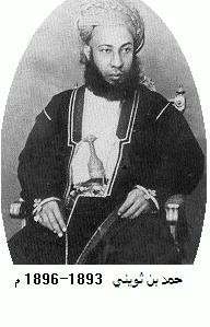 A black-and-white photograph of a man with a dark beard wearing a turban, a dark jacket, and a white shirt, sitting, and looking at the viewer