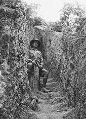 A man in military uniform sitting down in a trench. On both sides are walls of dirt, with sandbags on top.