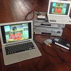 A laptop with the game footage sits atop two VHS players. Wires connect them to another laptop nearby, with a VHS cartridge to the right.