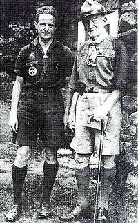 two men outdoors, both wearing Scout uniforms: short sleeve shirt, shorts and knee socks, the older man on the right wearing a campaign hat and holding a cane