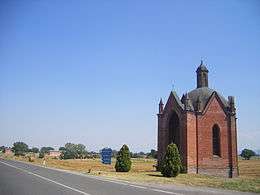 Photo of a small brick chapel on a highway amid rolling pastures. A sign reads Voghera to the right and Piacenza to the left.