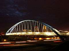 A large arched suspension bridge passes over a busy highway.  The nighttime skyline is seen through the cable supports.
