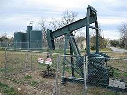 Photo shows a dark gray oil pump jack from a few yards away. A cylindrical oil tank is in the background.