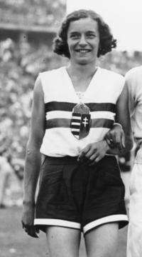 A black and white photograph of a female athlete with short cut hair. She wears a white sleeveless top with two horizontal stripes and a crest in the middle of her chest, and dark shorts.