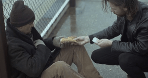 A screenshot from the music video; Browne's character is giving a homeless man (portrayed by Carter) food and taking back his money he offered him first. The video has low colouring and is set on a dreary day, puddles are still present from the rain.