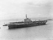 Black and white photograph showing a World War II-era aircraft carrier in a body of water which is surrounded by low hills. The bow of a smaller ship is visible behind the aircraft carrier.